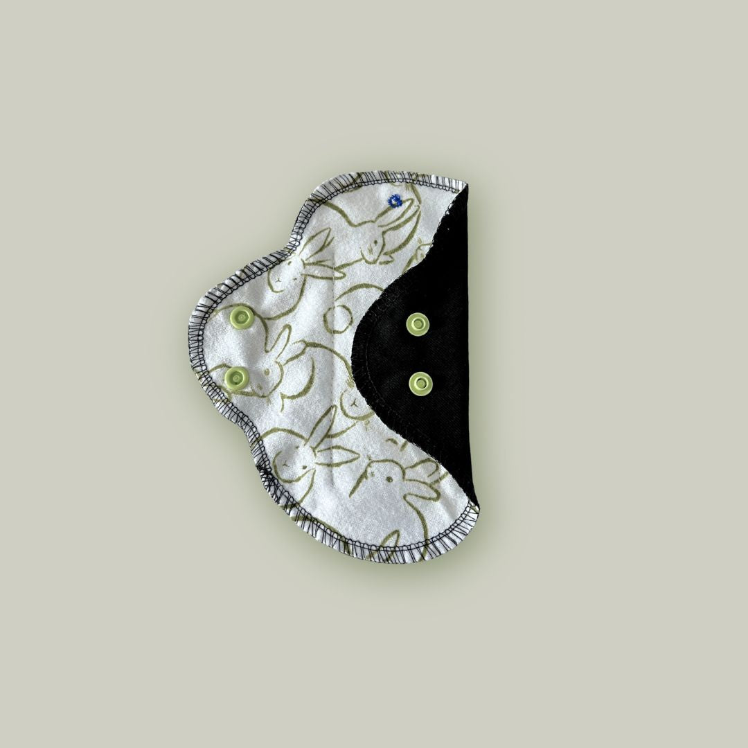 Teen reusable pad in bunny print partially folded to show both patterned fabric side and black absorbent layer with green snaps.