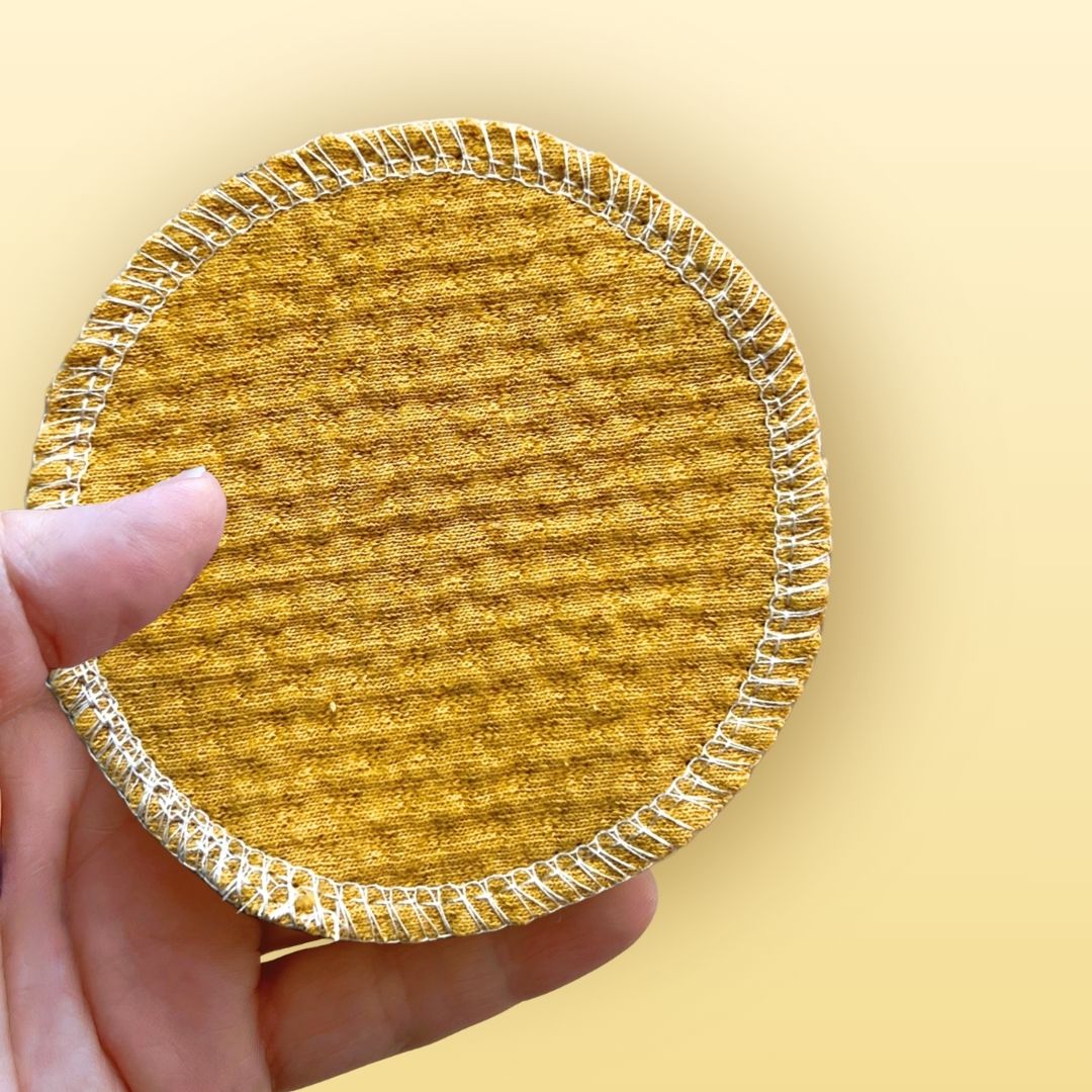 Hand holding a single 4-inch organic cotton face pad in warm golden sand color