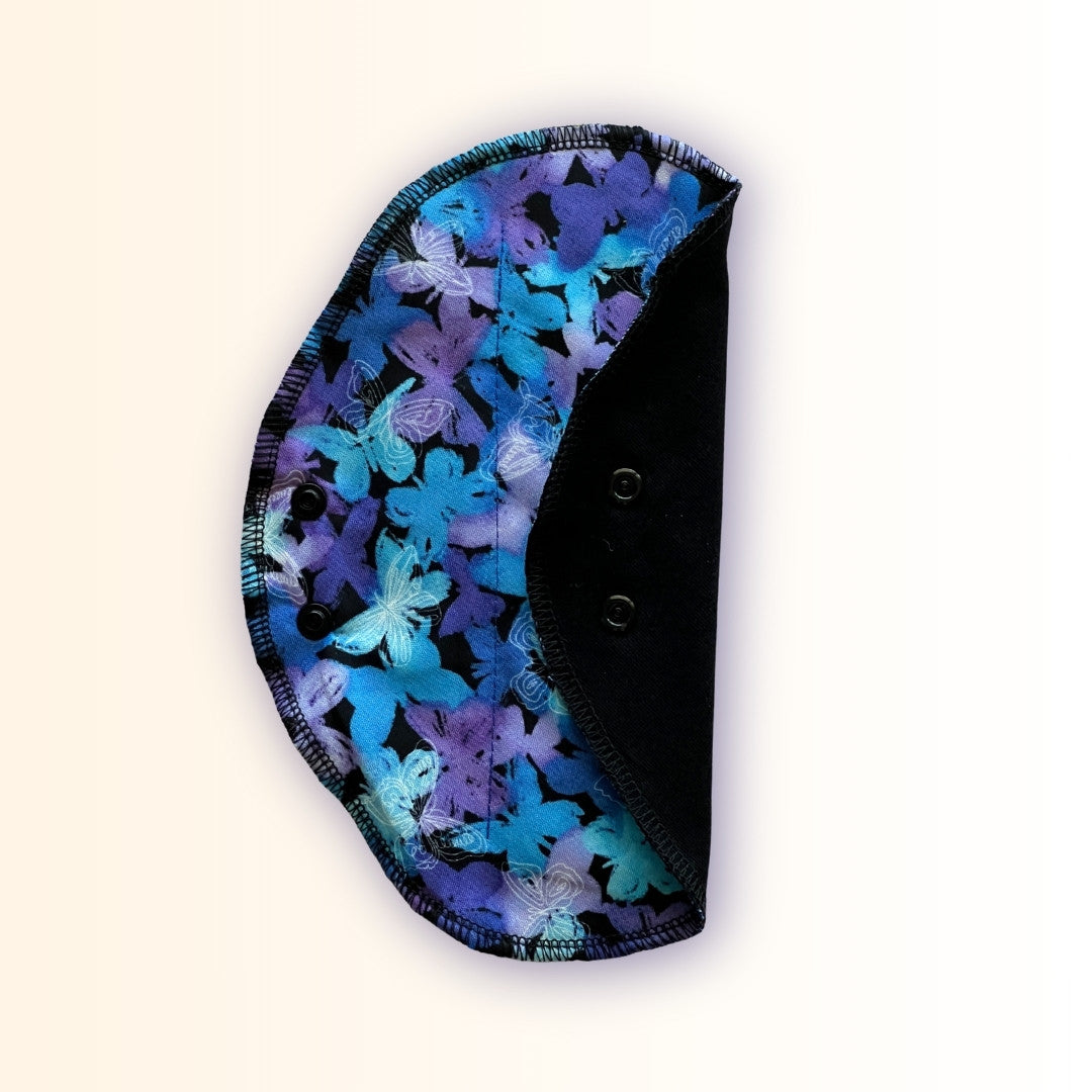 Blue butterfly reusable period pad folded to show the teal and purple butterfly print, black cotton absorbent side, and snap wings from Hume’s Period Care.