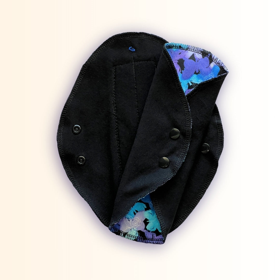Blue Butterfly Reusable Period Pad shown open and folded to display the black cotton absorbent side, snap wings, and butterfly print from Hume’s Period Care.