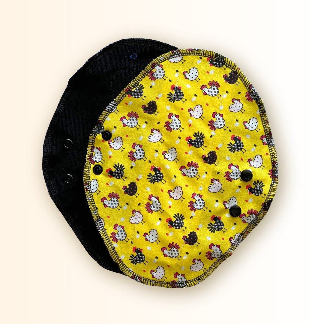 Yellow chicken reusable period pad shown open with black cotton absorbent side, snap wings, and cheerful chicken print from Hume’s Period Care.