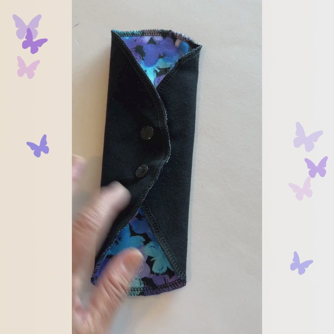 Video showing the Blue Butterfly Reusable Period Pad from Hume’s Period Care with teal and purple butterfly print, black cotton absorbent side, snap wings, and washable cloth pad design.
