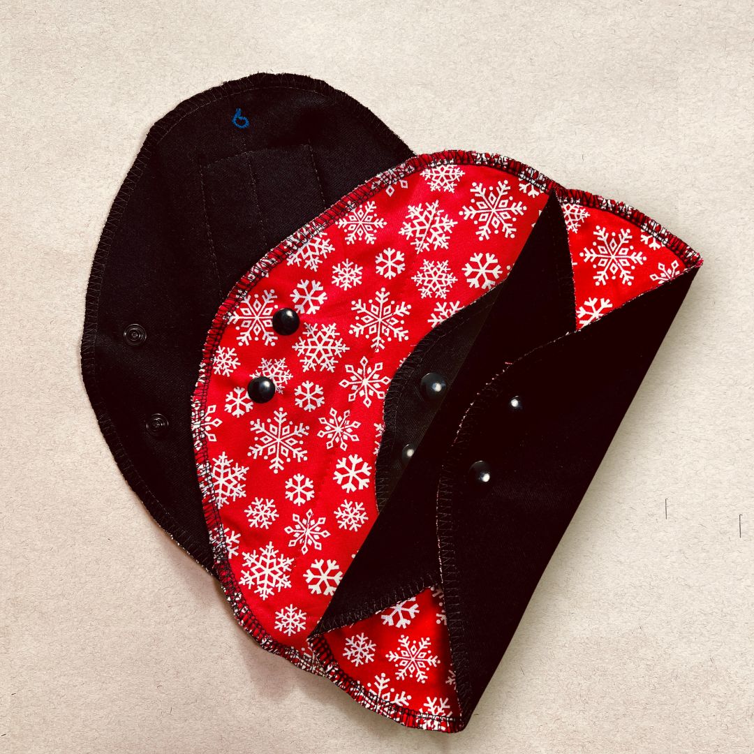 Black reusable menstrual pad with Christmas print — organic cotton core, holiday edition. showing one pad flat over black fabric absorbent side and one half folded and one pad folded snapped