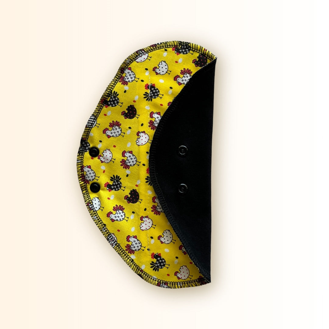 Yellow chicken reusable period pad folded to show the bright chicken print, black cotton absorbent side, and snap wings from Hume’s Period Care.