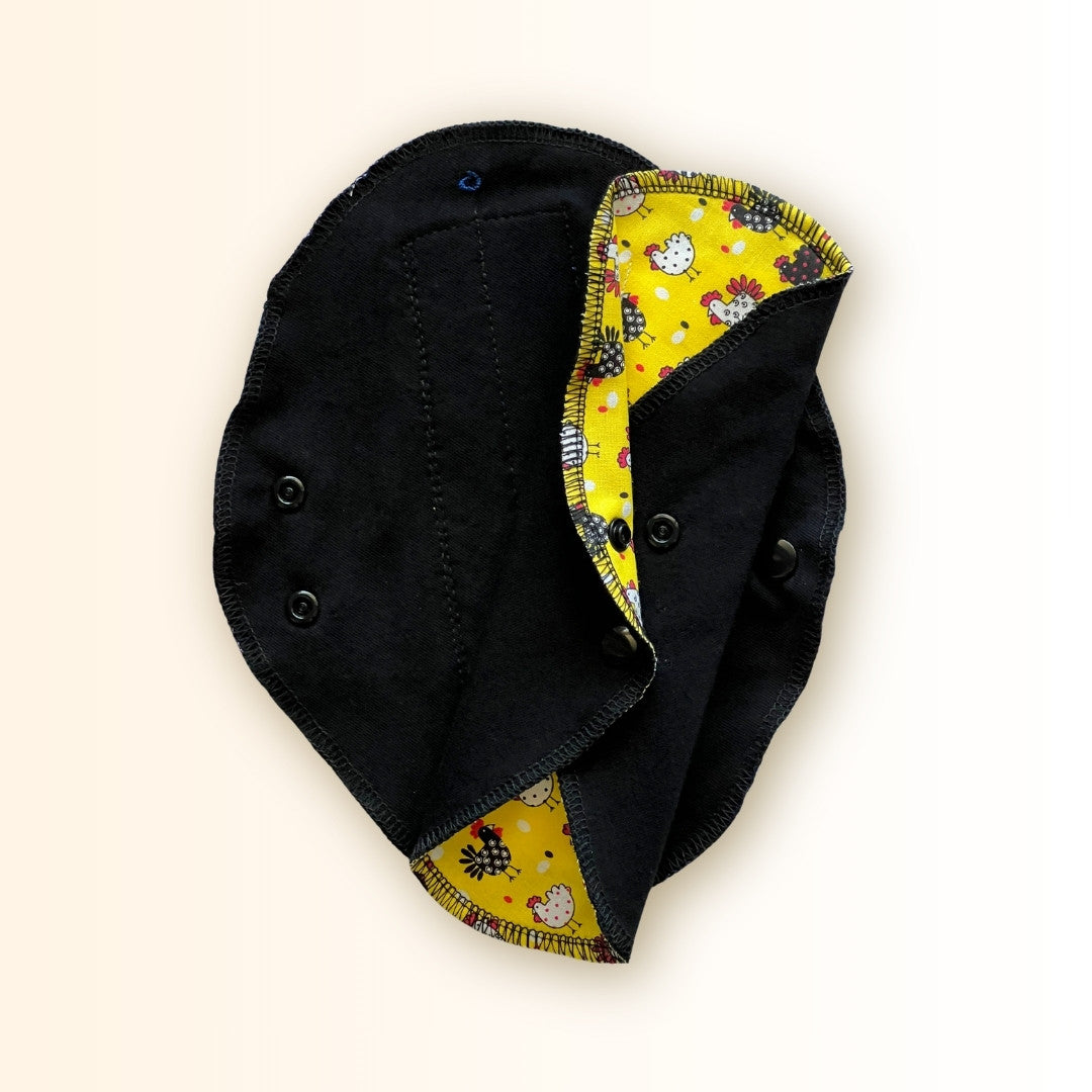 Yellow Chicken Reusable Period Pad shown open and folded to display the yellow chicken print, black cotton side, and snap closure from Hume’s Period Care.
